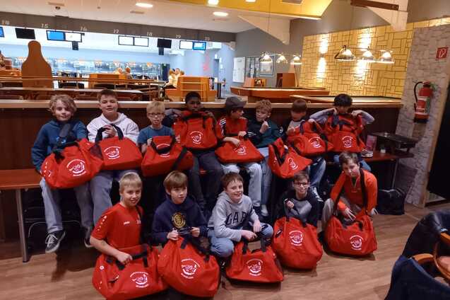 Gruppe von Jungen sitzt mit roten Sporttaschen in einem Bowling-Caf&eacute;, im Hintergrund sind Bowlingbahnen sichtbar.