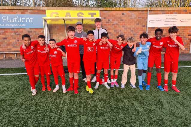 Gruppe von Jugendlichen in roten Fu&szlig;balltrikots, posierend mit Trainer auf einem Kunstrasenfeld.