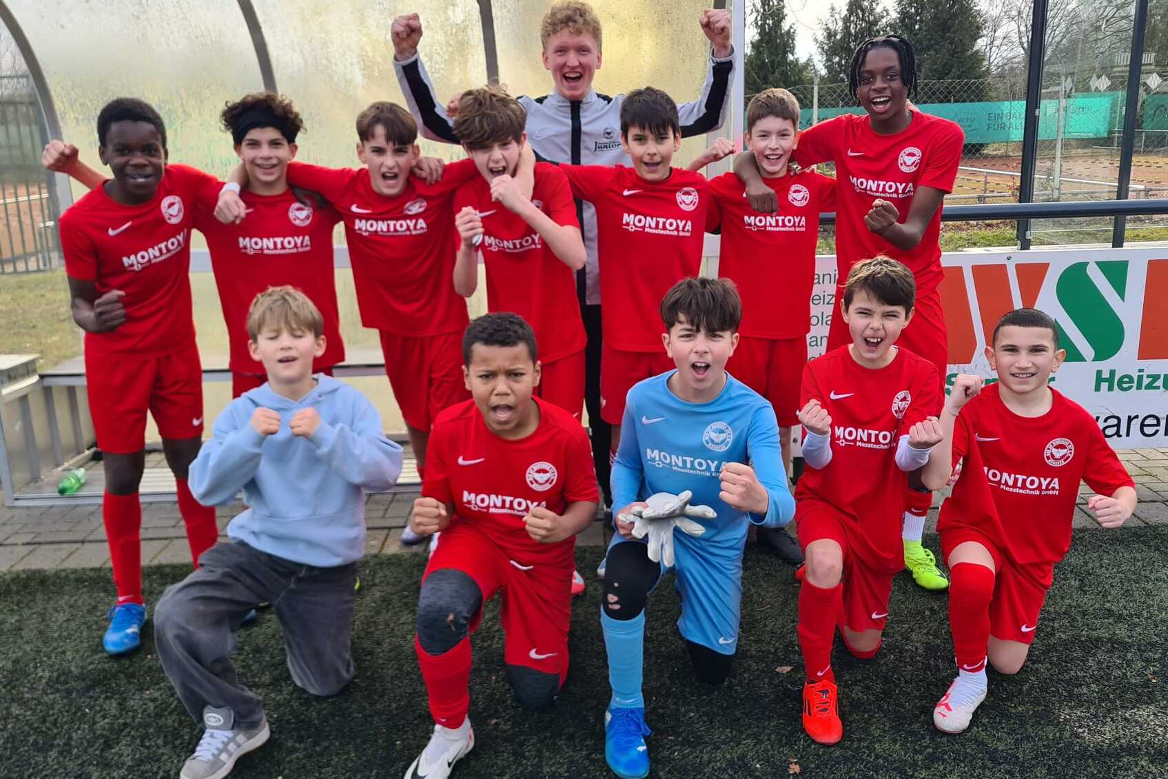 Gruppe junger Fu&szlig;ballspieler in roten Trikots posiert jubelnd auf einem Sportplatz, ein Torwart in blauer Uniform vorne.