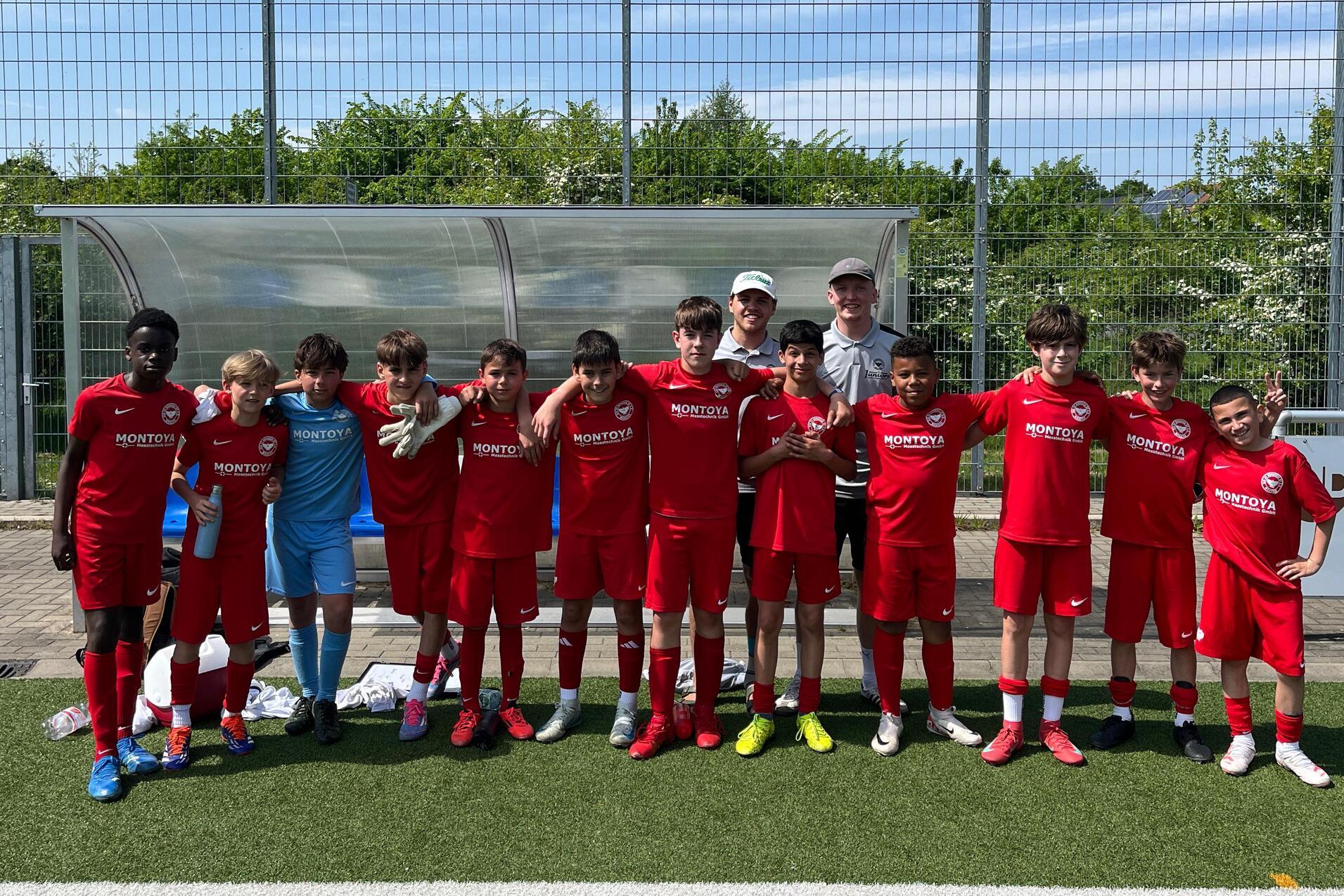 Gruppe von elf Fu&szlig;ballspielern in roten Trikots, posierend auf einem Sportplatz mit einem Trainer im Hintergrund.