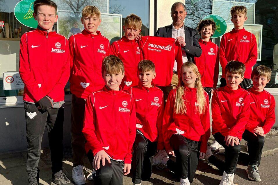 Gruppe von zehn jungen Sportlern in roten Trainingsanz&uuml;gen, posierend vor einem Fenster mit einem Erwachsenen.