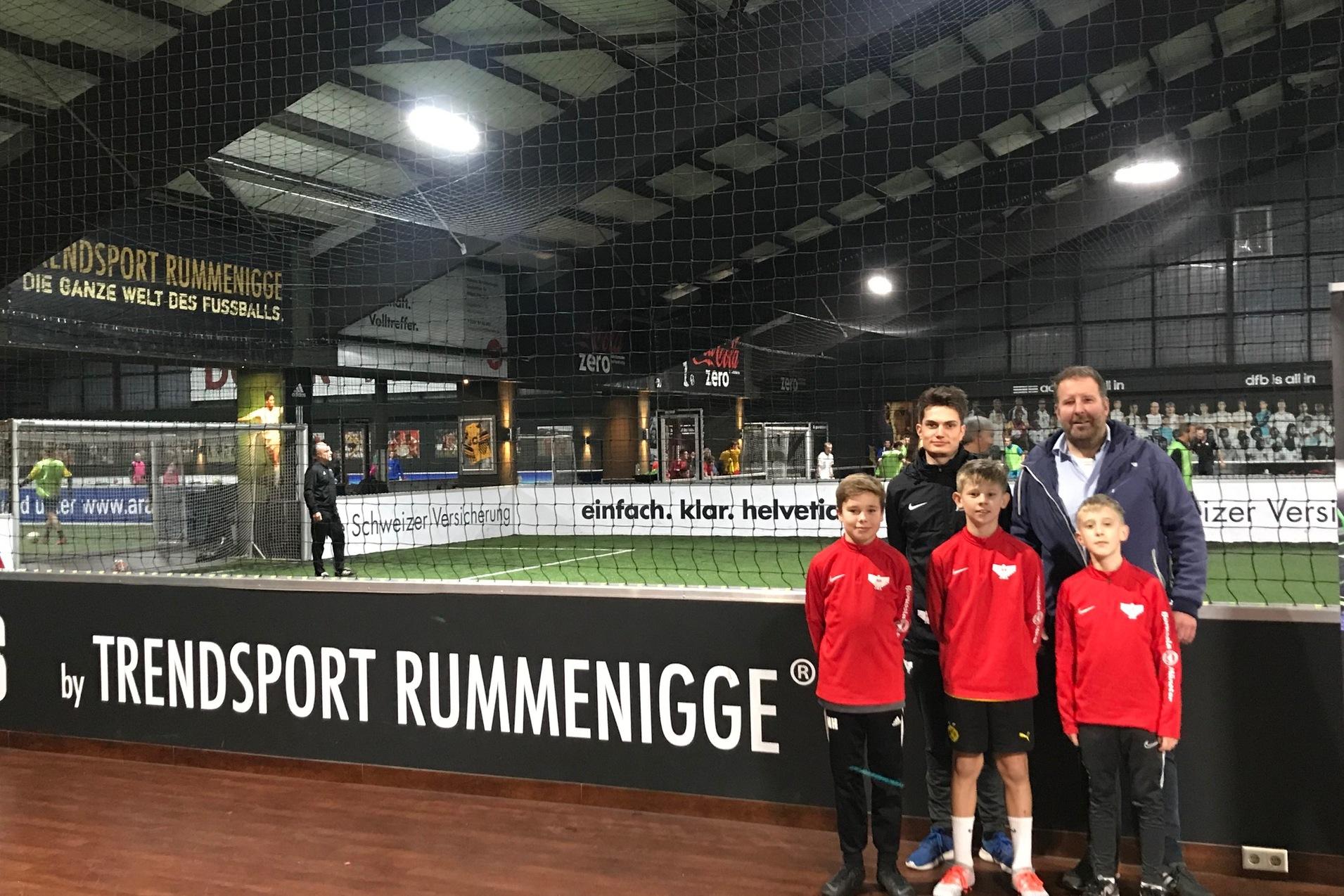 Gruppe von vier Jungs in roten Sportjacken posiert mit einem Erwachsenen in einer Soccer-Halle vor einem Fu&szlig;ballfeld.