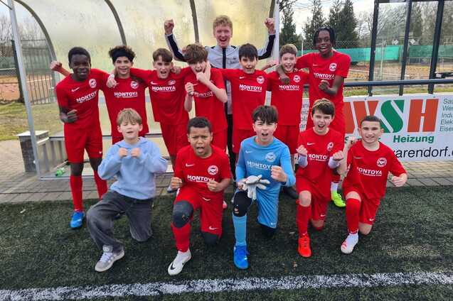 Gruppe junger Fu&szlig;ballspieler in roten Trikots posiert jubelnd auf einem Sportplatz, ein Torwart in blauer Uniform vorne.
