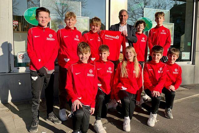 Gruppe von zehn jungen Sportlern in roten Trainingsanz&uuml;gen, posierend vor einem Fenster mit einem Erwachsenen.