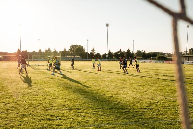 Fu&szlig;balltraining auf gr&uuml;nem Rasenplatz bei Sonnenuntergang, Sicht durch ein Tornetz auf die Spieler.