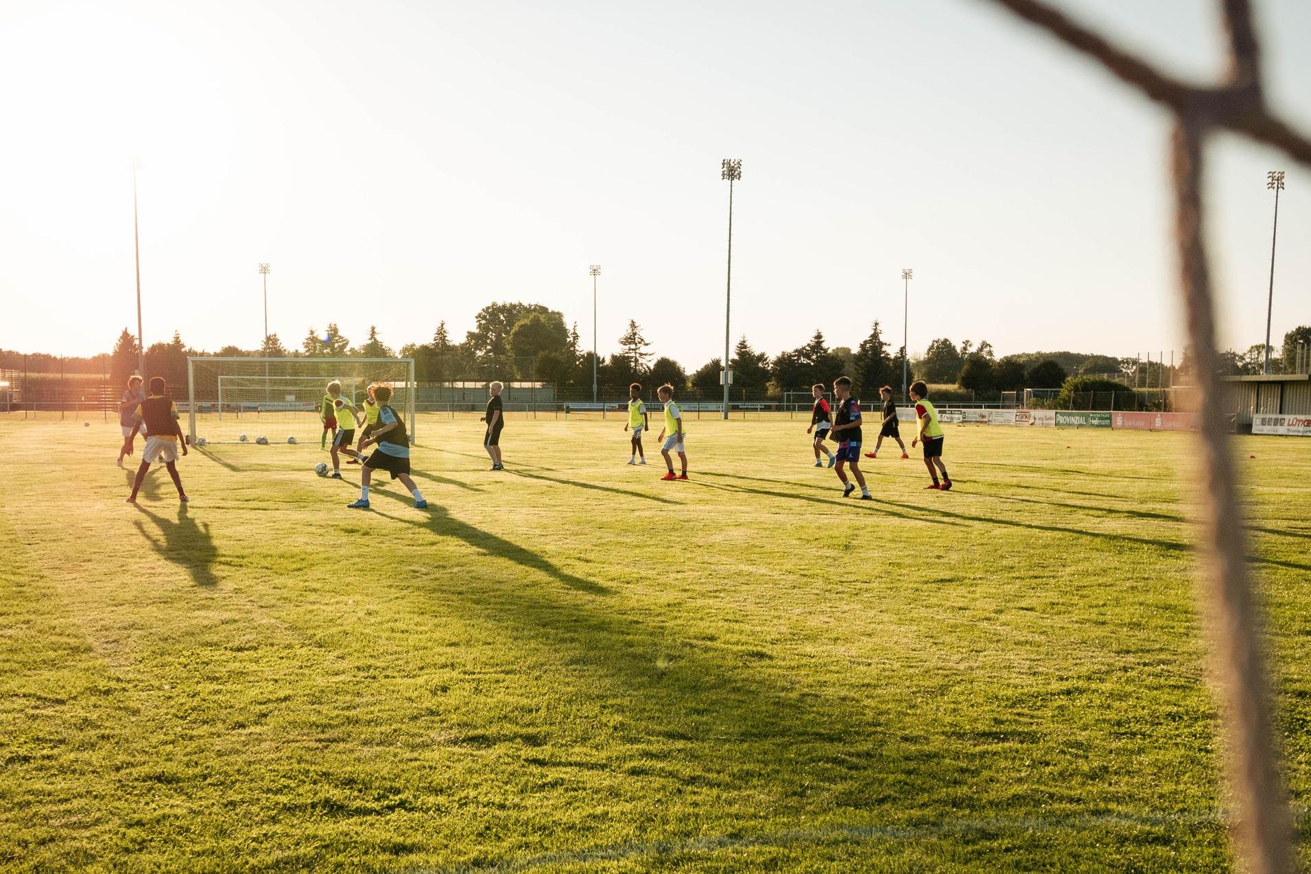 Fu&szlig;balltraining auf gr&uuml;nem Rasenplatz bei Sonnenuntergang, Sicht durch ein Tornetz auf die Spieler.