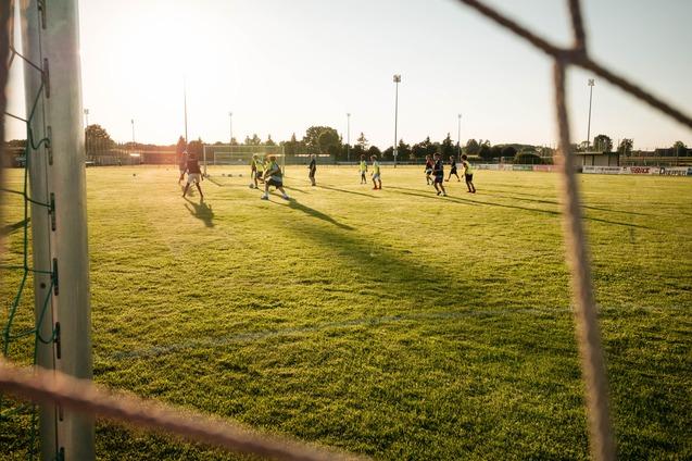 Fu&szlig;balltraining auf gr&uuml;nem Rasenplatz bei Sonnenuntergang, Sicht durch ein Tornetz auf die Spieler.