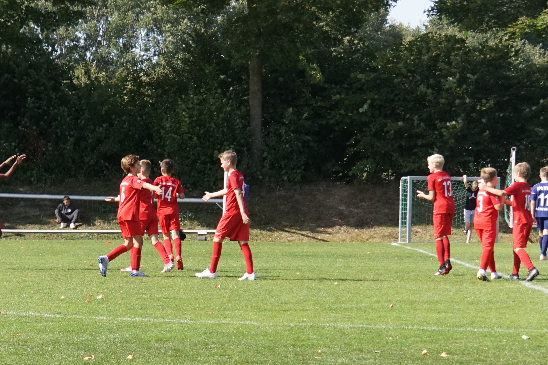 Spieler in roten Trikots feiern auf einem Fu&szlig;ballfeld, im Hintergrund sind Trainer und Zuschauer zu sehen.