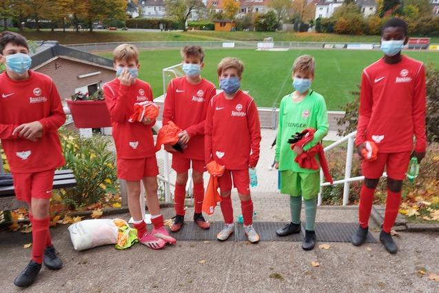 Sechs Kinder in roten Fu&szlig;balltrikots und Masken stehen vor einem Sportplatz mit herbstlichem Laub.