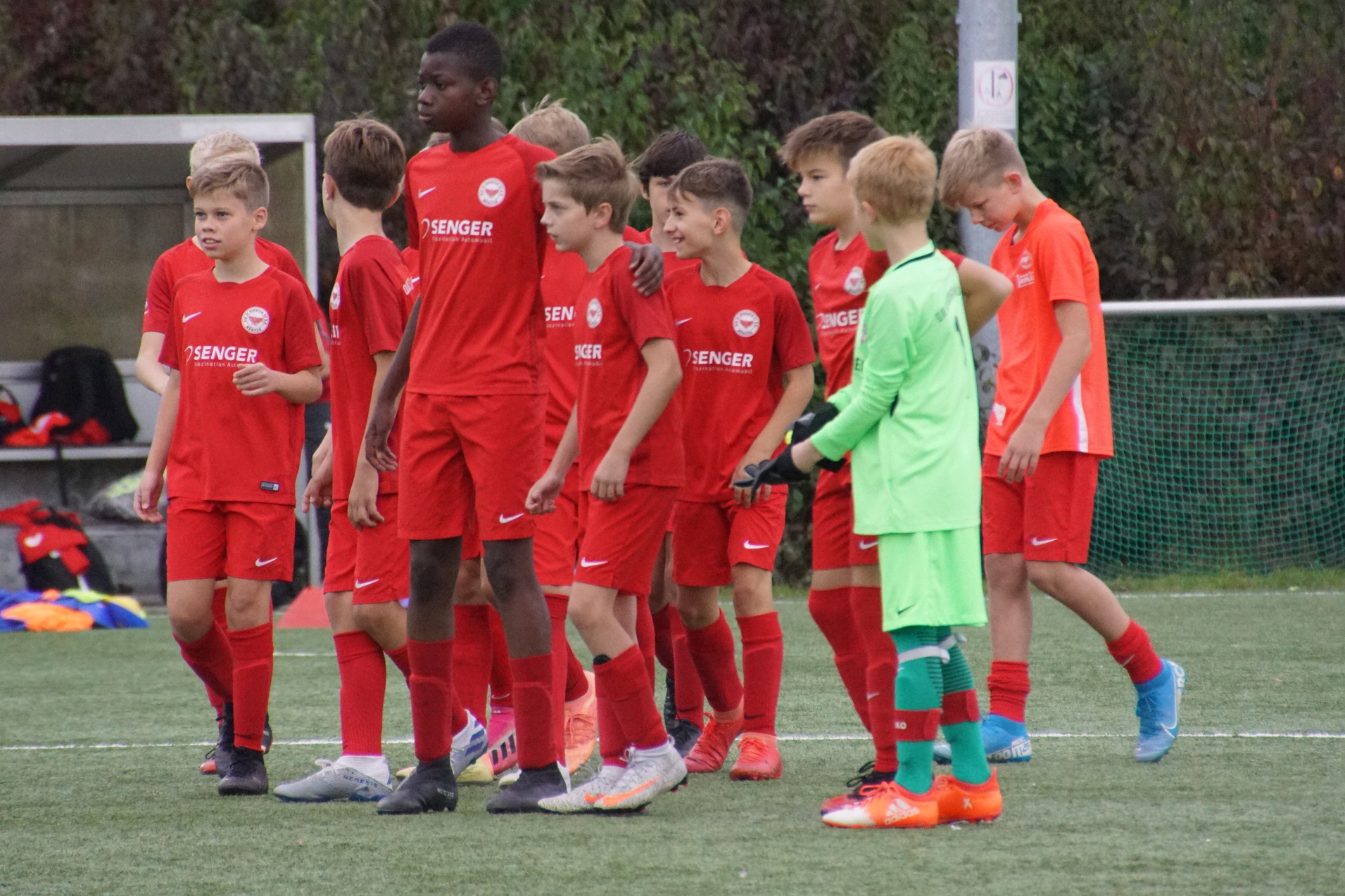 Gruppe von Jungen in roten Fu&szlig;balltrikots auf einem Spielfeld, vorbereitet auf ein Spiel, mit einem Torwart in gr&uuml;ner Ausr&uuml;stung.
