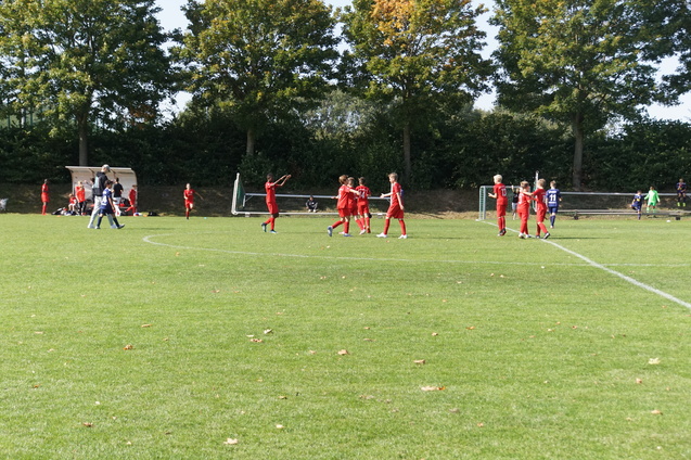 Spieler in roten Trikots feiern auf einem Fu&szlig;ballfeld, im Hintergrund sind Trainer und Zuschauer zu sehen.