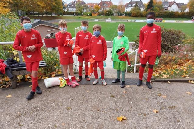 Sechs Kinder in roten Fu&szlig;balltrikots und Masken stehen vor einem Sportplatz mit herbstlichem Laub.