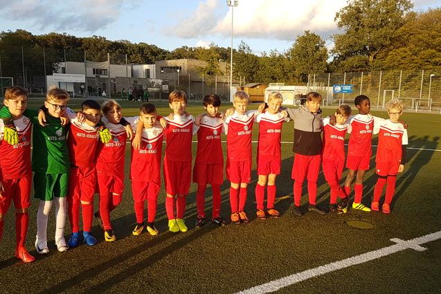 Gruppe von zehn jungen Fu&szlig;ballspielern in roten Trikots, auf einem Fu&szlig;ballfeld stehend, mit Sportplatz im Hintergrund.