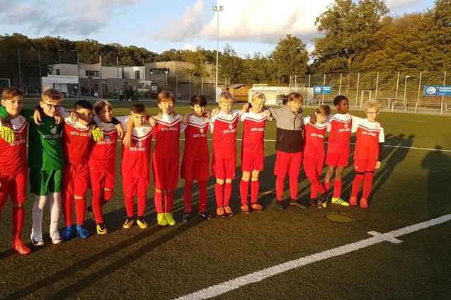 Gruppe von zehn jungen Fu&szlig;ballspielern in roten Trikots, auf einem Fu&szlig;ballfeld stehend, mit Sportplatz im Hintergrund.