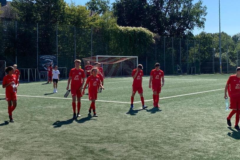 Gruppe von Spielern in roten Trikots auf einem Fu&szlig;ballplatz unter blauem Himmel mit B&auml;umen im Hintergrund.