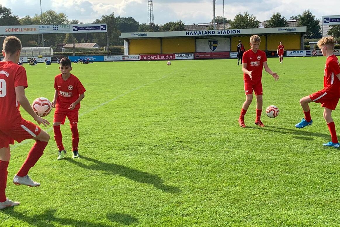Vier Kinder in roten Fu&szlig;balltrikots &uuml;ben mit B&auml;llen auf einem gr&uuml;nen Rasenplatz unter leicht bew&ouml;lktem Himmel.