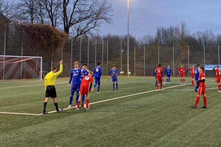 Spielszene auf einem Fu&szlig;ballfeld mit Spielern in roten und blauen Trikots; Schiedsrichter zeigt auf eine Regelverletzung.