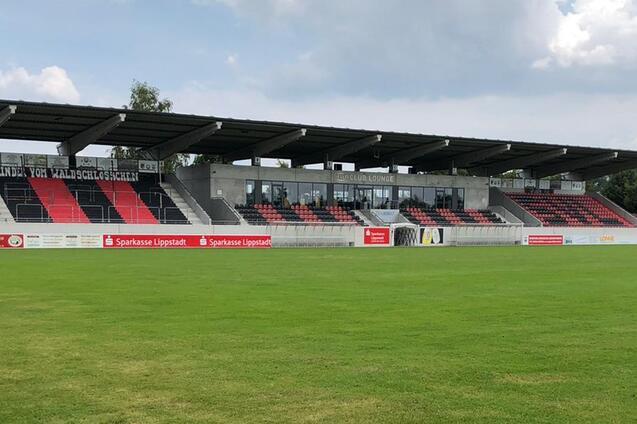 Blick auf ein Fu&szlig;ballfeld mit gr&uuml;nem Rasen und Trib&uuml;ne im Hintergrund unter einem teils bew&ouml;lkten Himmel.