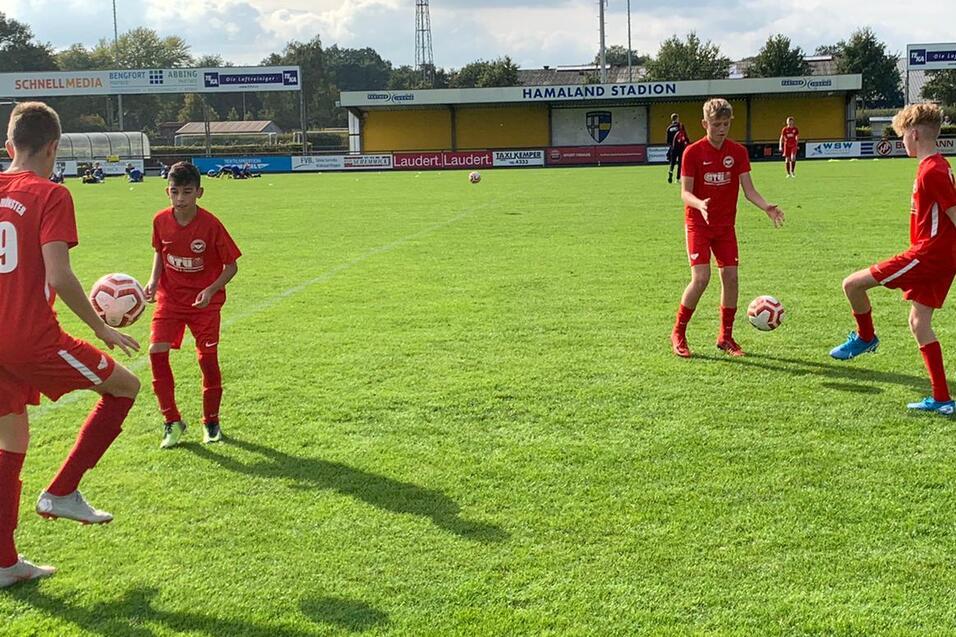 Vier Kinder in roten Fu&szlig;balltrikots &uuml;ben mit B&auml;llen auf einem gr&uuml;nen Rasenplatz unter leicht bew&ouml;lktem Himmel.