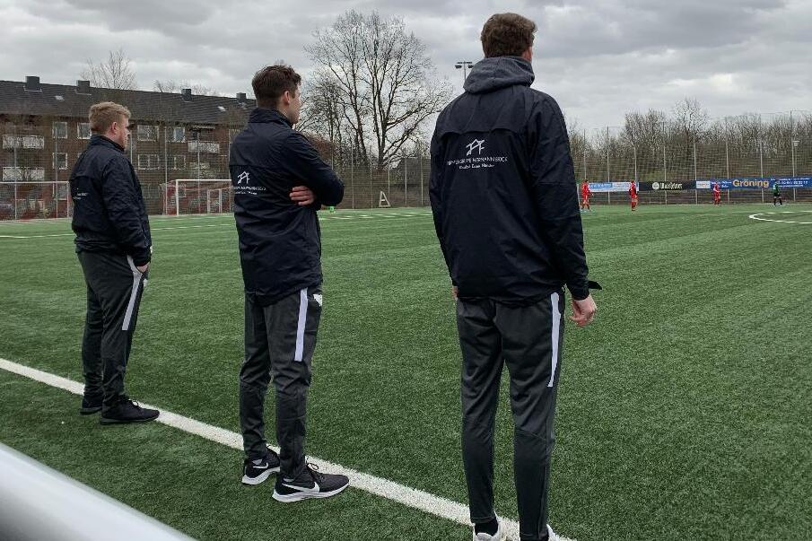 Drei Trainer in schwarzen Jacken beobachten ein Fu&szlig;ballspiel auf einem Kunstrasenplatz unter bew&ouml;lktem Himmel.