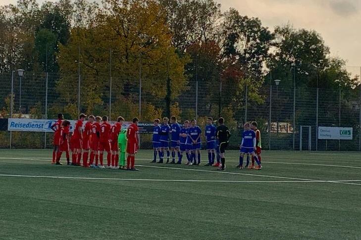 Zwei Fu&szlig;ballmannschaften in roten und blauen Trikots stehen auf einem Kunstrasenfeld zusammen, um sich zu besprechen.