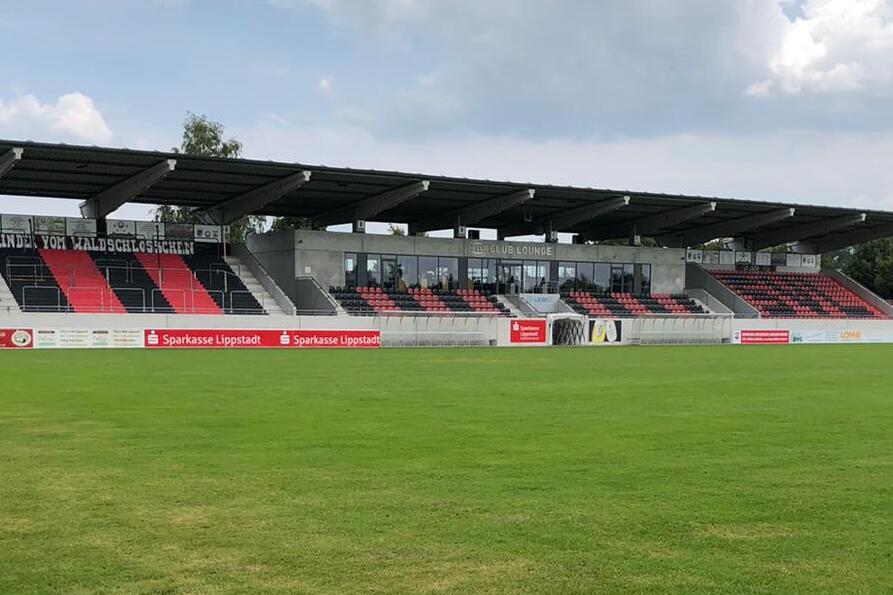 Blick auf ein Fu&szlig;ballfeld mit gr&uuml;nem Rasen und Trib&uuml;ne im Hintergrund unter einem teils bew&ouml;lkten Himmel.