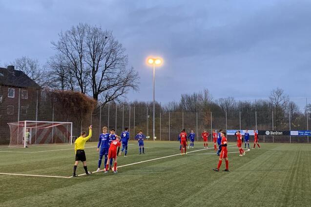 Spielszene auf einem Fu&szlig;ballfeld mit Spielern in roten und blauen Trikots; Schiedsrichter zeigt auf eine Regelverletzung.