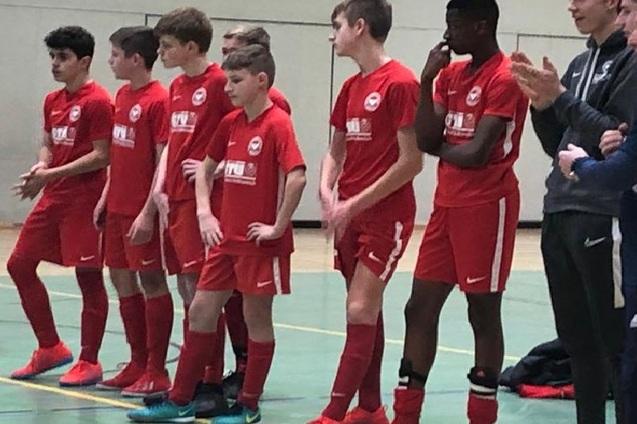 Eine Gruppe von sieben Jugendlichen in roten Fu&szlig;balltrikots steht an der Seitenlinie eines Hallenfu&szlig;ballspiels.