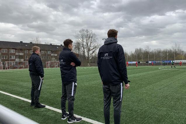 Drei Trainer in schwarzen Jacken beobachten ein Fu&szlig;ballspiel auf einem Kunstrasenplatz unter bew&ouml;lktem Himmel.