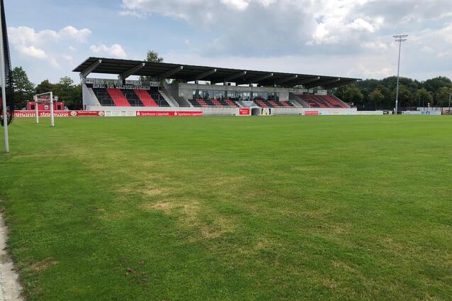 Blick auf ein Fu&szlig;ballfeld mit gr&uuml;nem Rasen und Trib&uuml;ne im Hintergrund unter einem teils bew&ouml;lkten Himmel.
