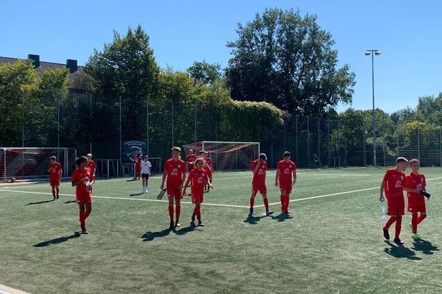 Gruppe von Spielern in roten Trikots auf einem Fu&szlig;ballplatz unter blauem Himmel mit B&auml;umen im Hintergrund.