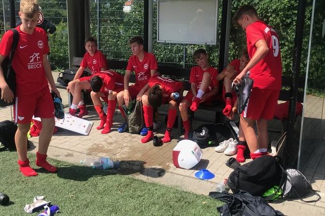 Gruppe von Jugendlichen in roten Sporttrikots sitzt auf einer Bank, einige ziehen Fu&szlig;ballschuhe an oder entspannen.