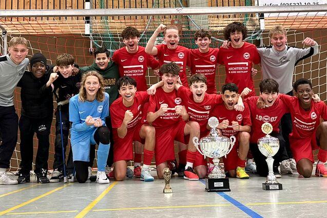 Gruppe von Jugendlichen in roten Trikots mit Pokalen vor einem Fu&szlig;balltor in einer Sporthalle.