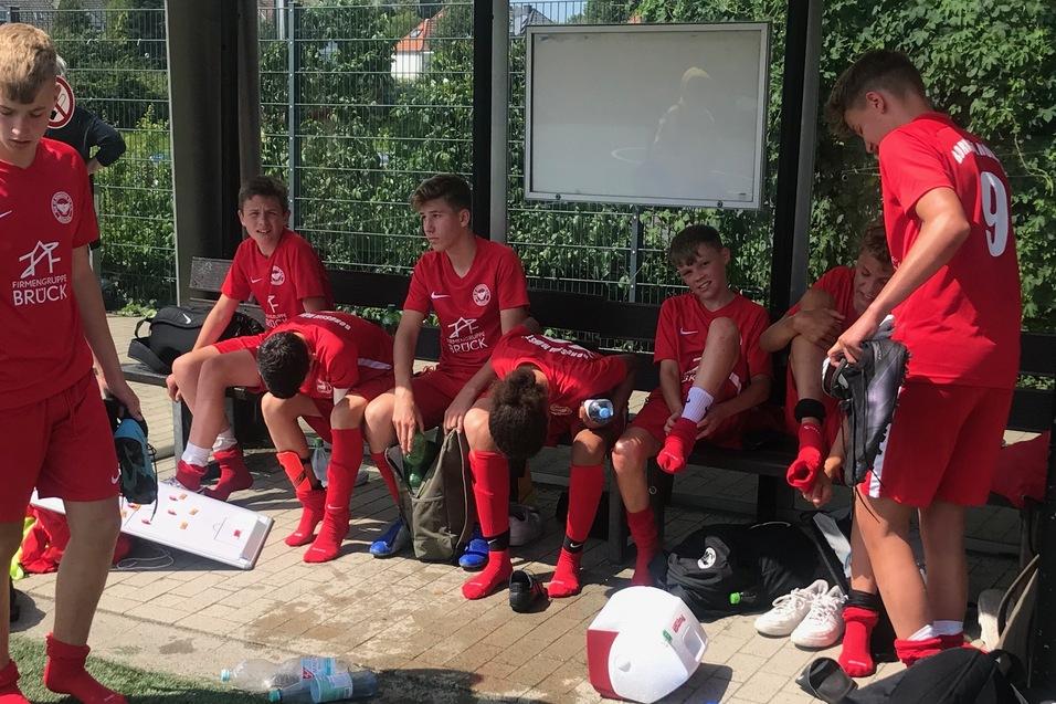 Gruppe von Jugendlichen in roten Sporttrikots sitzt auf einer Bank, einige ziehen Fu&szlig;ballschuhe an oder entspannen.