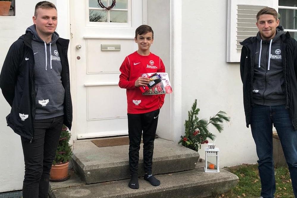 Drei Personen stehen vor einem Haus: Ein Junge in rotem Hoodie h&auml;lt ein Geschenk, flankiert von zwei Erwachsenen.