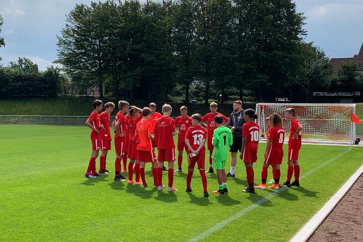 Eine Gruppe junger Fu&szlig;ballspieler in roten Trikots versammelt sich auf einem Rasenplatz mit einem Tor im Hintergrund.