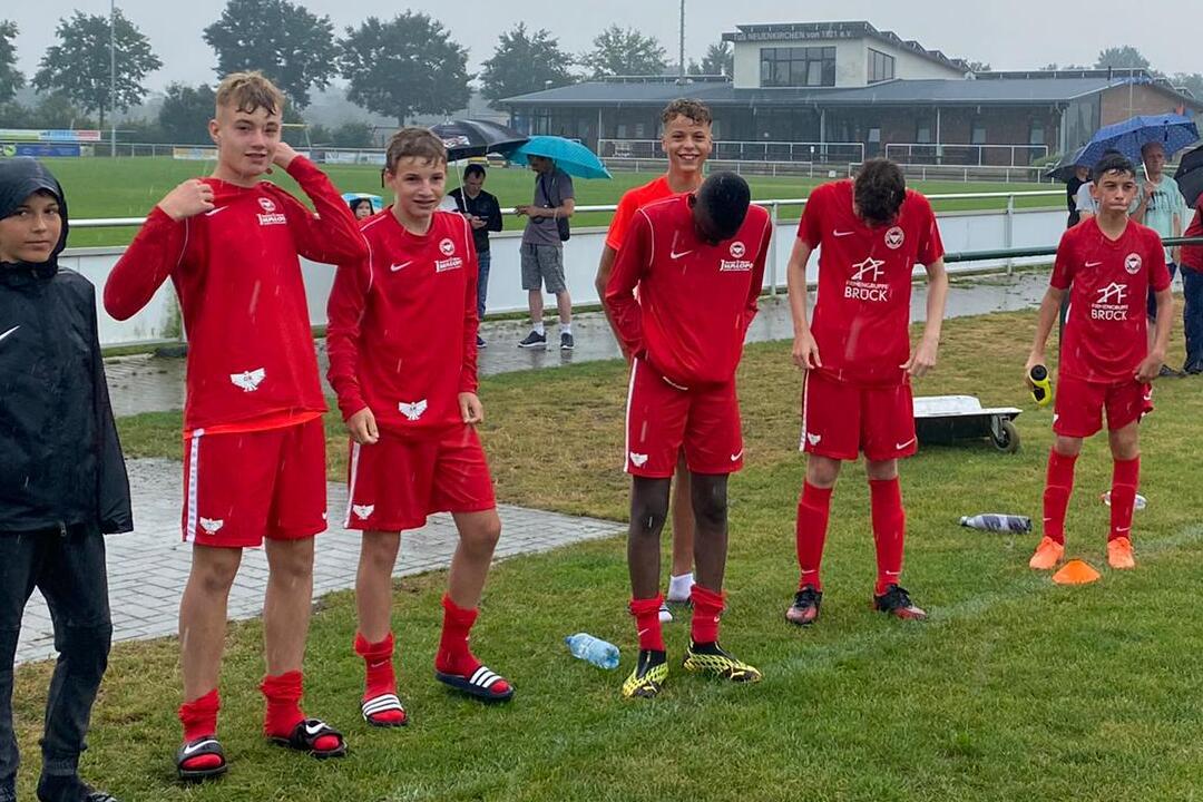 Sechs Jugendliche in roten Fu&szlig;balltrikots stehen bei Regen an einem Sportplatz, einige halten Regenschirme.