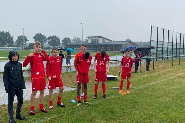 Sechs Jugendliche in roten Fu&szlig;balltrikots stehen bei Regen an einem Sportplatz, einige halten Regenschirme.
