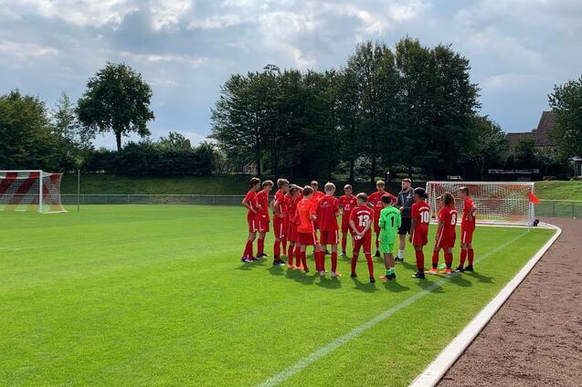 Eine Gruppe junger Fu&szlig;ballspieler in roten Trikots versammelt sich auf einem Rasenplatz mit einem Tor im Hintergrund.