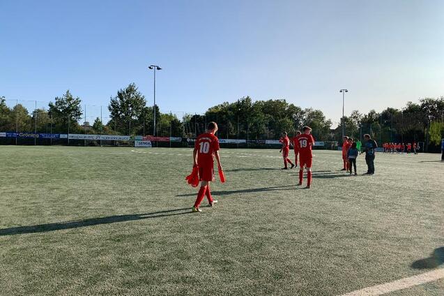 Spieler in roten Trikots verlassen den Fu&szlig;ballplatz, umgeben von B&auml;umen und einem klaren blauen Himmel.