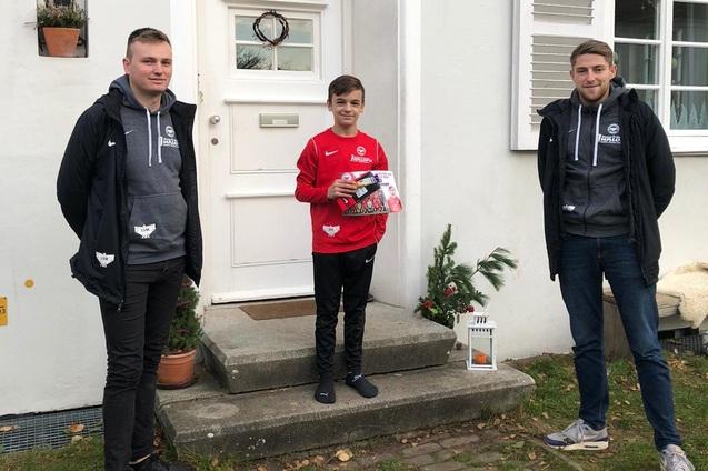 Drei Personen stehen vor einem Haus: Ein Junge in rotem Hoodie h&auml;lt ein Geschenk, flankiert von zwei Erwachsenen.