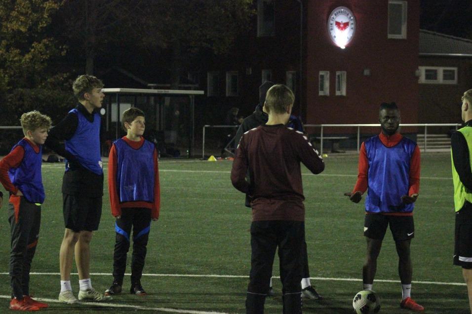 Gruppentraining von sieben Fu&szlig;ballspielern in blauen und roten Wams auf einem Kunstrasenplatz bei schwachem Licht.