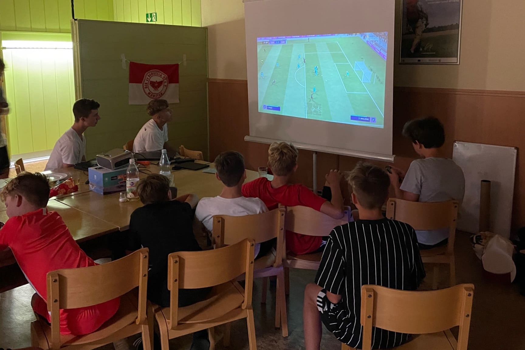 Gruppe von Kindern sitzt auf St&uuml;hlen und schaut ein Fu&szlig;ballspiel auf einer Leinwand.