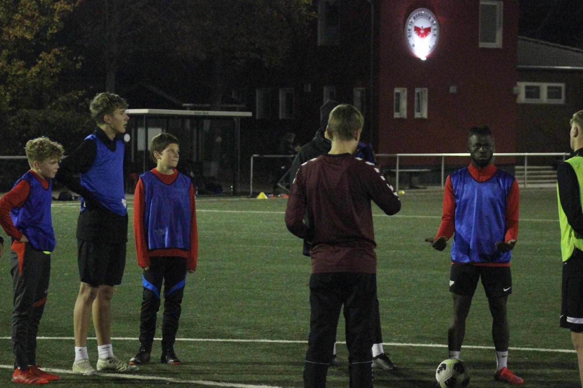 Gruppentraining von sieben Fu&szlig;ballspielern in blauen und roten Wams auf einem Kunstrasenplatz bei schwachem Licht.