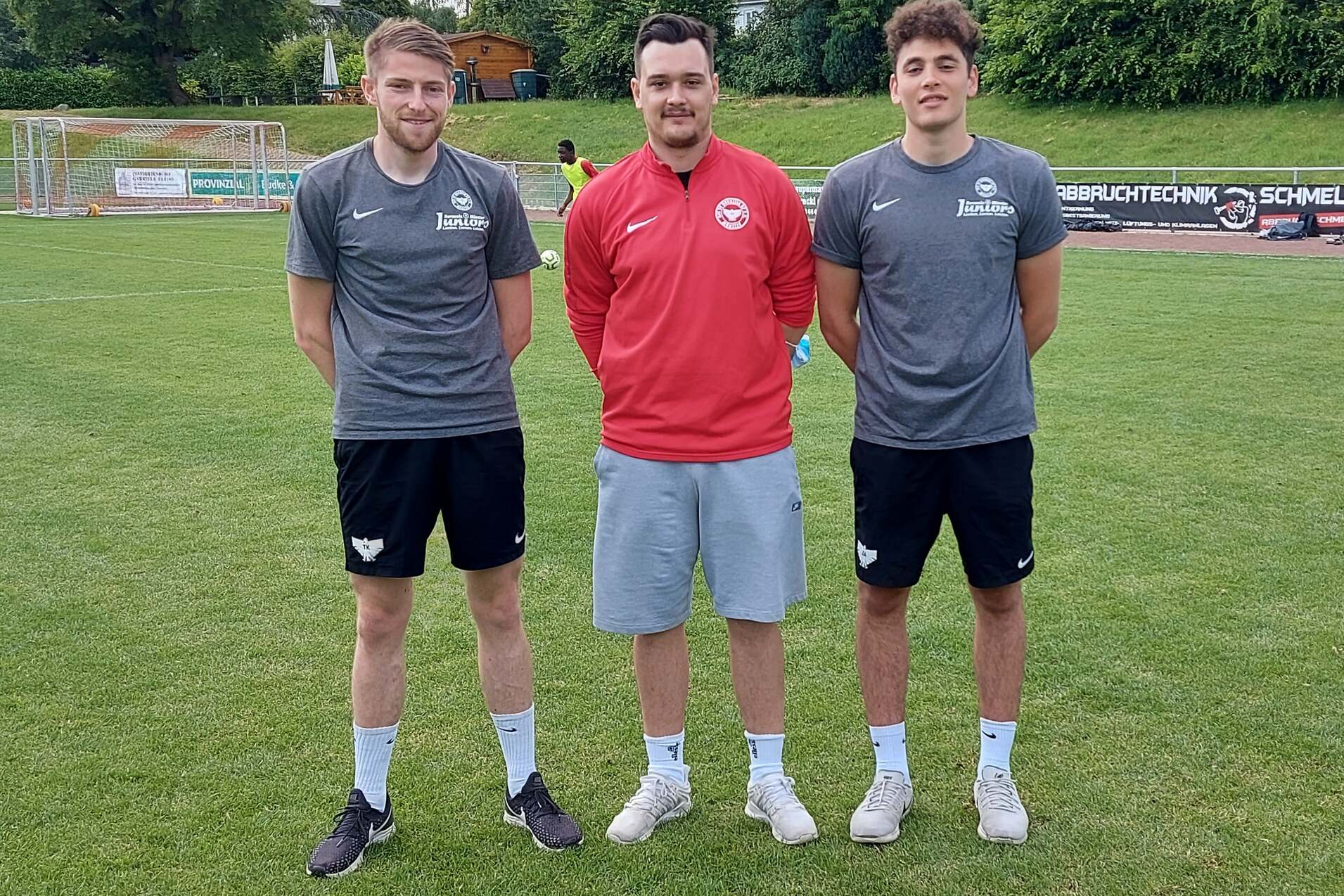 Drei Fu&szlig;ballspieler stehen auf einem Rasenplatz, umgeben von Werbebannern und B&auml;umen im Hintergrund.