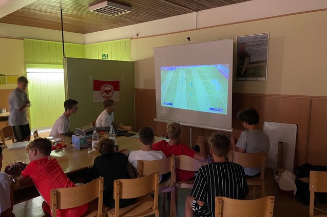 Gruppe von Kindern sitzt auf St&uuml;hlen und schaut ein Fu&szlig;ballspiel auf einer Leinwand.