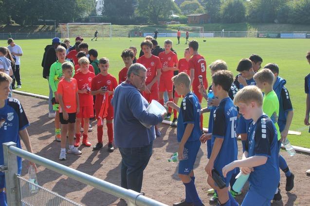 Gruppe von jungen Fu&szlig;ballspielern in verschiedenen Trikots versammelt sich um einen Trainer auf dem Sportplatz.