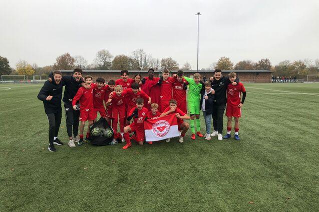 Gruppe von Jugendlichen in roten Fu&szlig;balltrikots posiert auf einem Kunstrasenplatz, h&auml;lt ein Banner.