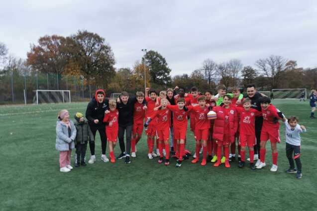Gruppe von Jugendlichen in roten Fu&szlig;balltrikots posiert auf einem Kunstrasenplatz, umgeben von B&auml;umen.