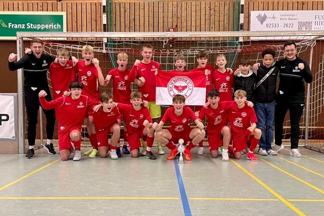 Gruppe von Jugendlichen in roten Trikots posiert jubelnd vor einem Handballtor in einer Sporthalle.